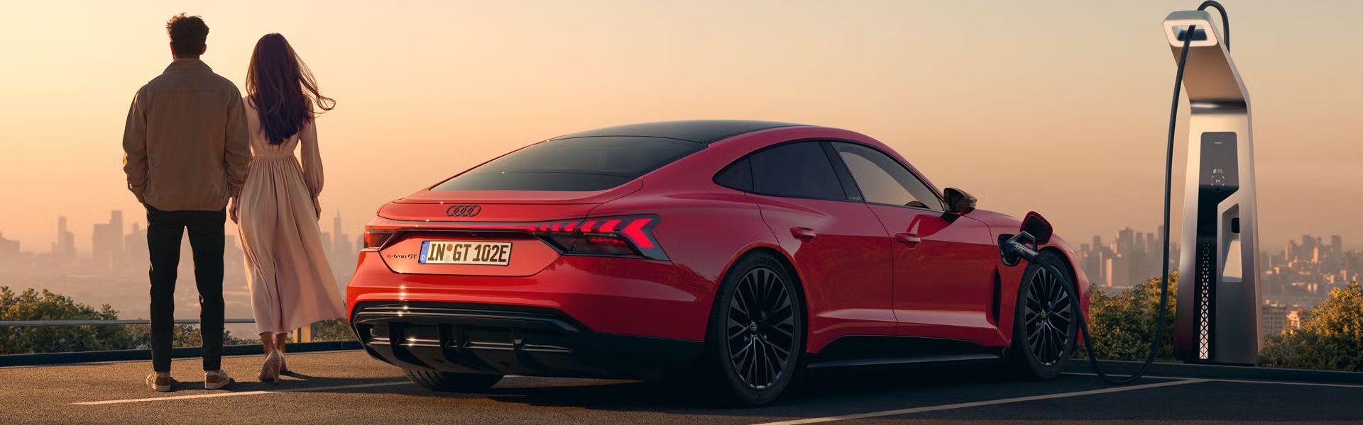 Ein roter Audi e-tron GT quattro steht auf einer Anhöhe über einer Stadt, die ins Licht eines Sonnenuntergangs gehüllt ist. Das Fahrzeug wird an einer öffentlicher Ladesäule geladen. Neben dem Fahrzeug stehen eine Frau und ein Mann.