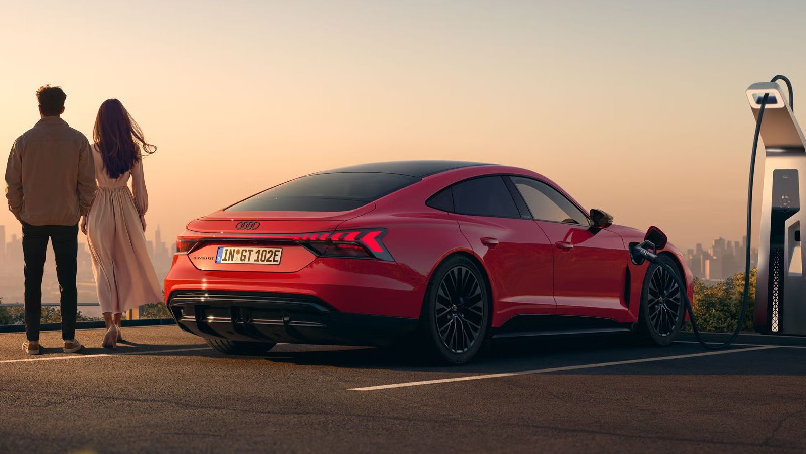 Ein roter Audi e-tron GT quattro steht auf einer Anhöhe über einer Stadt, die ins Licht eines Sonnenuntergangs gehüllt ist. Das Fahrzeug wird an einer öffentlicher Ladesäule geladen. Neben dem Fahrzeug stehen eine Frau und ein Mann.
