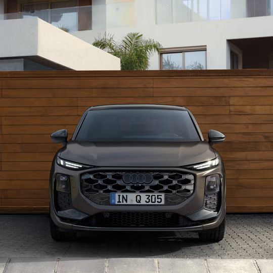 Der Q3 Sportback in Frontansicht parkt vor einem modernen Gebäude, dahinter eine Wand in Holzoptik.