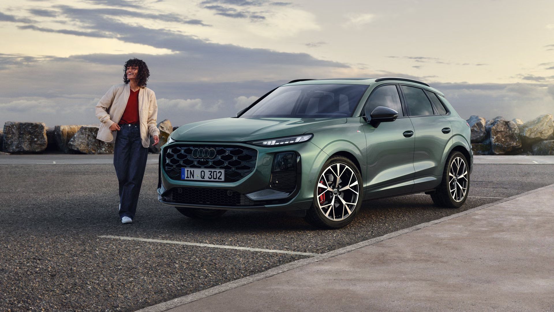 Seitliche, vordere Ansicht auf den geparkten Q3 SUV an einer Küstenstraße, daneben eine junge Frau. 