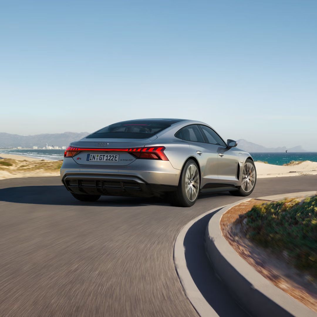 Ein silberner Audi S e-tron GT fährt auf einer kurvigen Küstenstraße mit Blick auf das Meer und eine Stadt im Hintergrund.