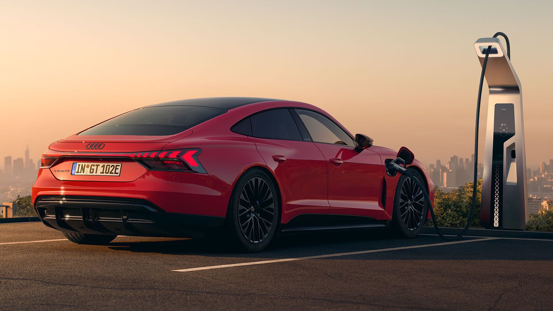 Ein roter Audi e-tron GT quattro steht auf einer Anhöhe über einer Stadt und wird an einer öffentlicher Ladesäule geladen.