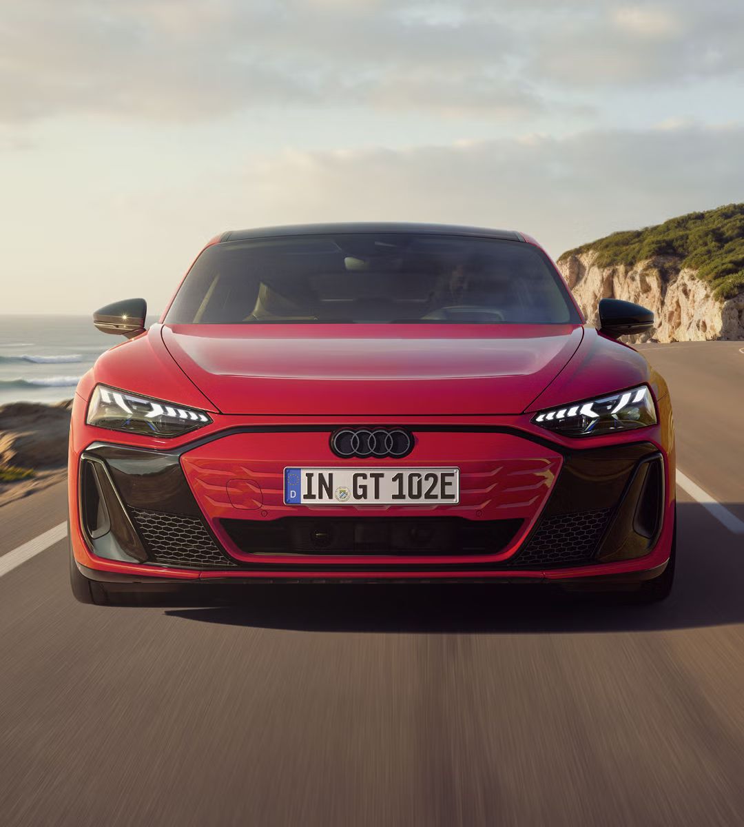 Frontansicht eines roten Audi e-tron GT quattro, der auf einer Küstenstraße fährt. Im Hintergrund sind Klippen, das Meer und der Himmel zu erkennen. 