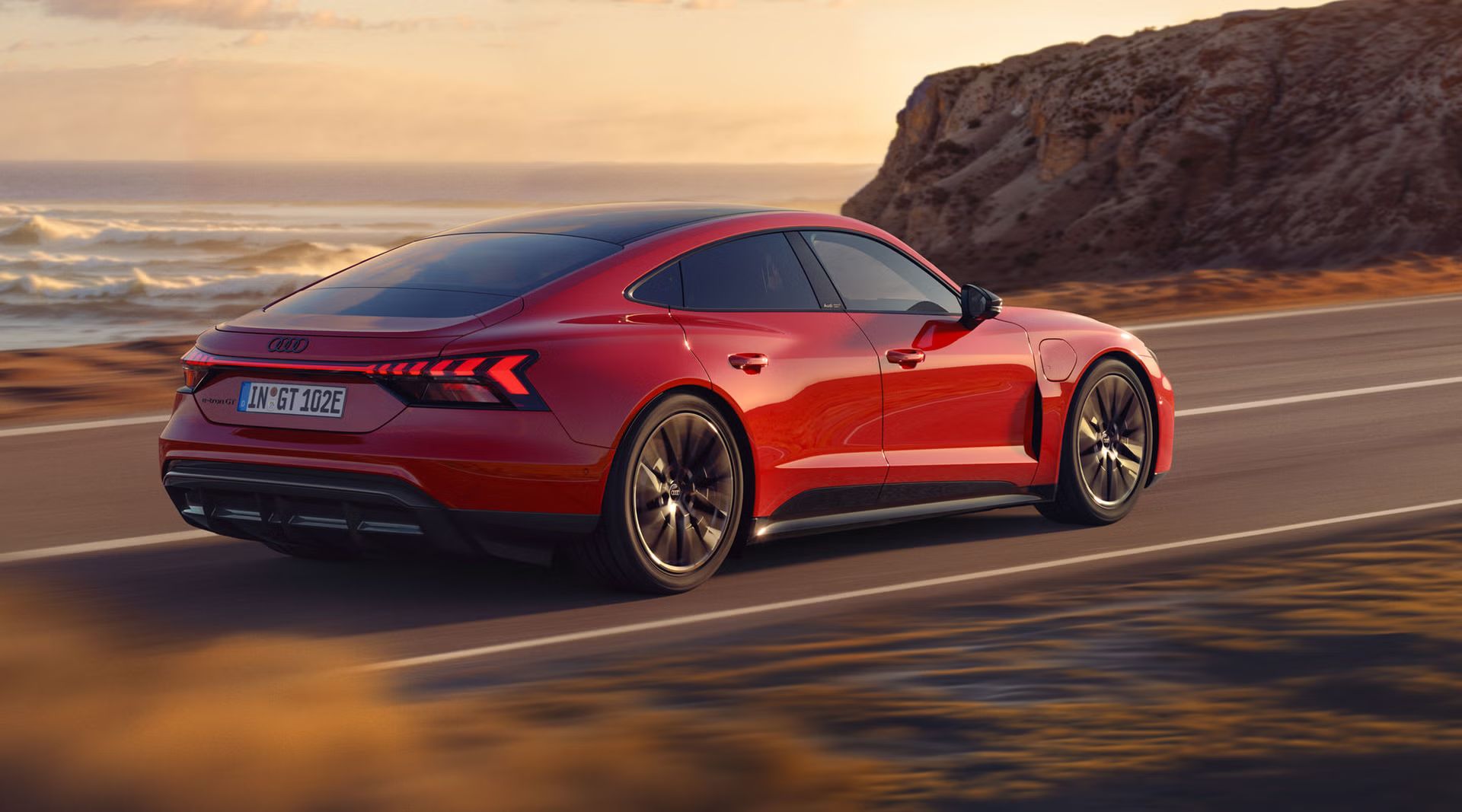 Dynamische Seitenansicht eines roten Audi e-tron GT quattro, der auf einer Küstenstraße fährt. Im Hintergrund sind Klippen, das Meer und der Himmel zu erkennen. Die gesamte Szenerie ist in das Licht eines Sonnenuntergangs gehüllt.